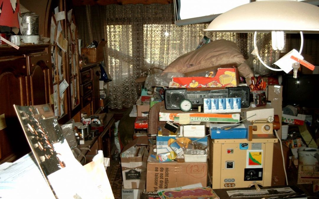 An untidy room filled with personal belonging piled up on the floor and surrounding furniture