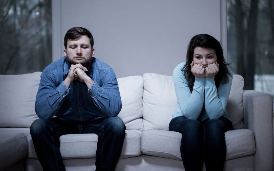 A young unhappy looking couple sat apart on a sofa looking at the floor