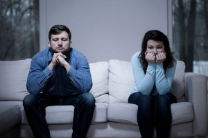 A young unhappy looking couple sat apart on a sofa looking at the floor