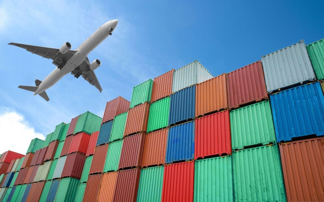A large cargo jet plane flying over a row of multi coloured stacked shipping containers