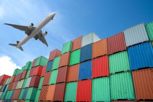 A large cargo jet plane flying over a row of multi coloured stacked shipping containers