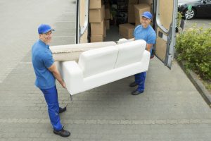 Two removals men unloading a sofa and rug from a delivery van filled with cardboard packing boxes