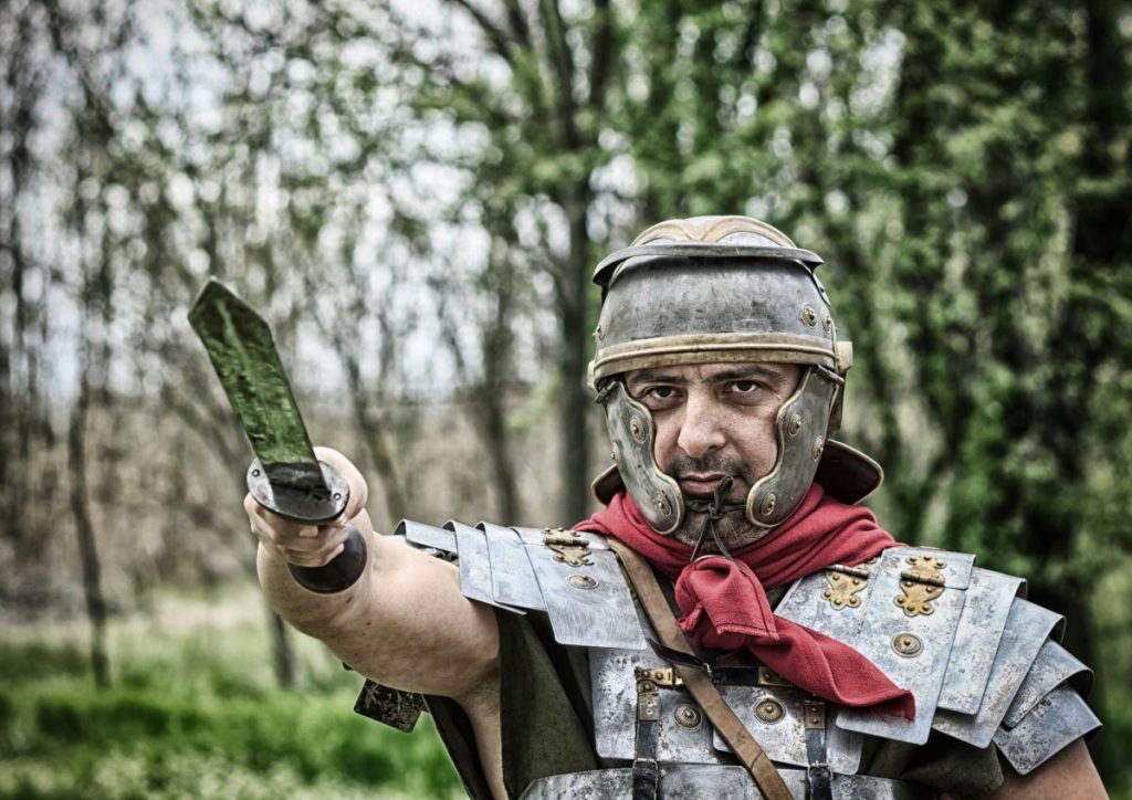 A man dressed in a Roman Gladiator outfit holding a sword aloft in front of a wood