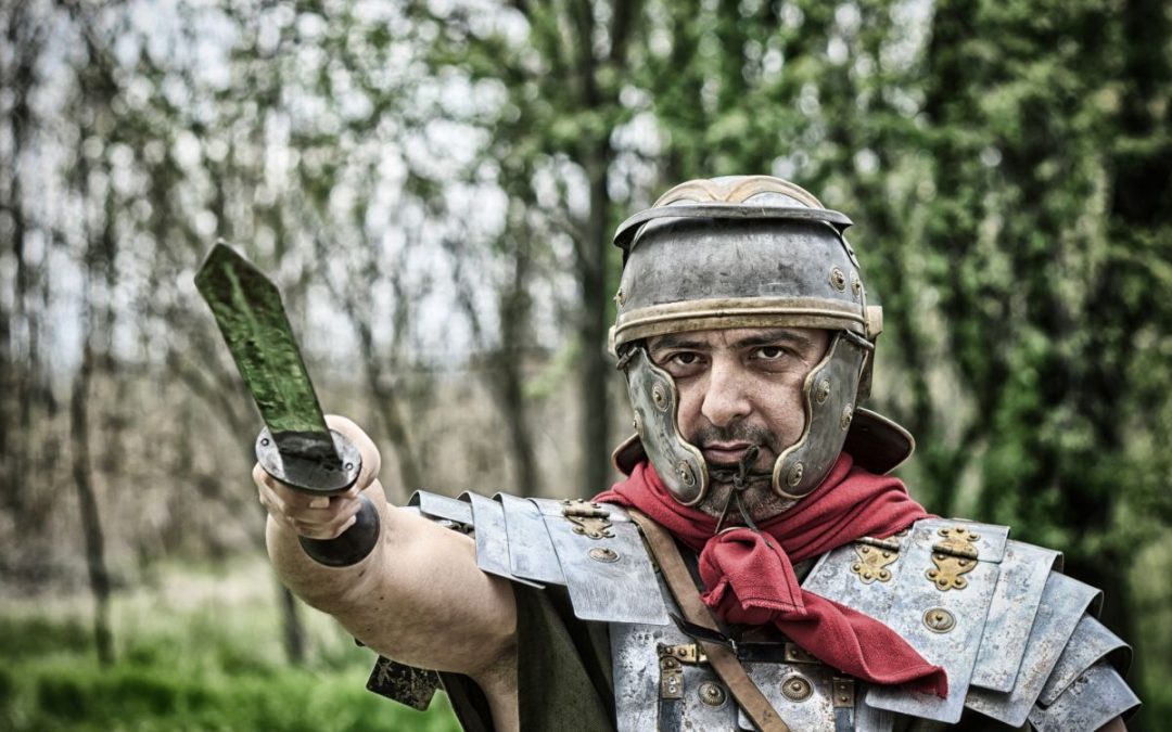 A man dressed in a Roman Gladiator outfit holding a sword aloft in front of a wood