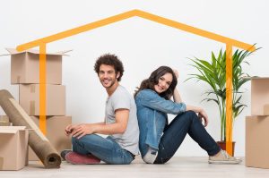 A happy couple sitting on the floor back to back surrounded by cardboard packing boxes, house plant, rug and a house shaped line sitting over them