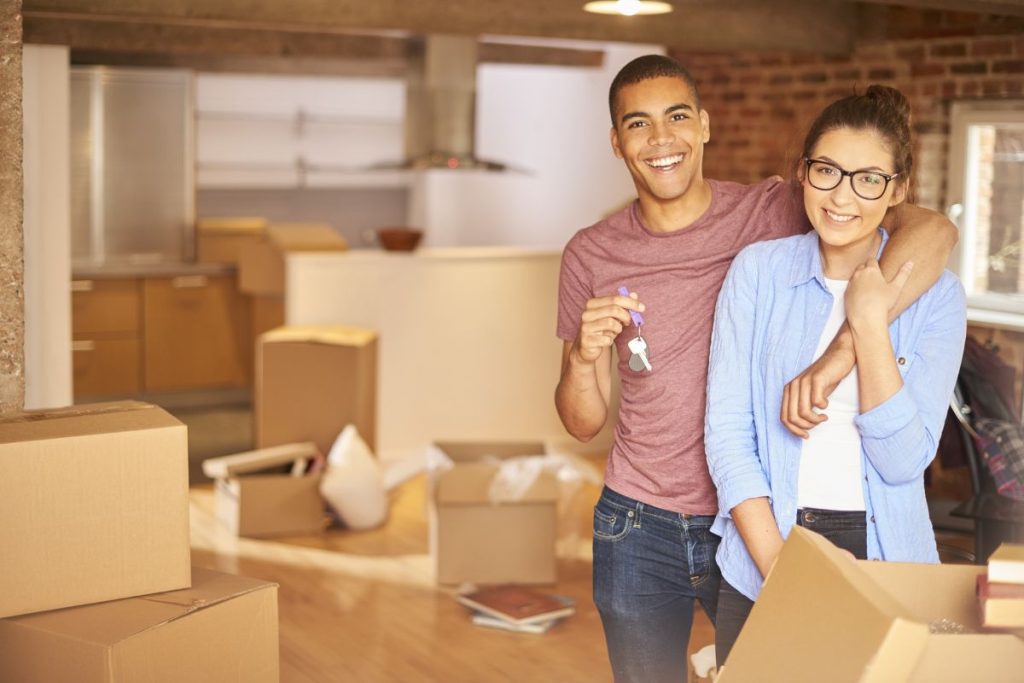 A smiling young couple with keys to their new home, surrounds by lots of partially unpacked removals boxes