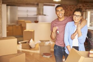 A smiling young couple with keys to their new home, surrounds by lots of partially unpacked removals boxes