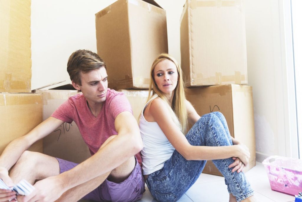 An unhappy looking couple sitting back to back surrounded by packed cardboard boxes