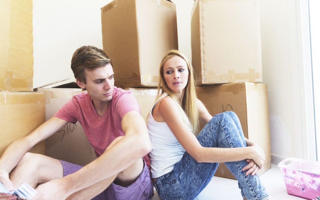 An unhappy looking couple sitting back to back surrounded by packed cardboard boxes