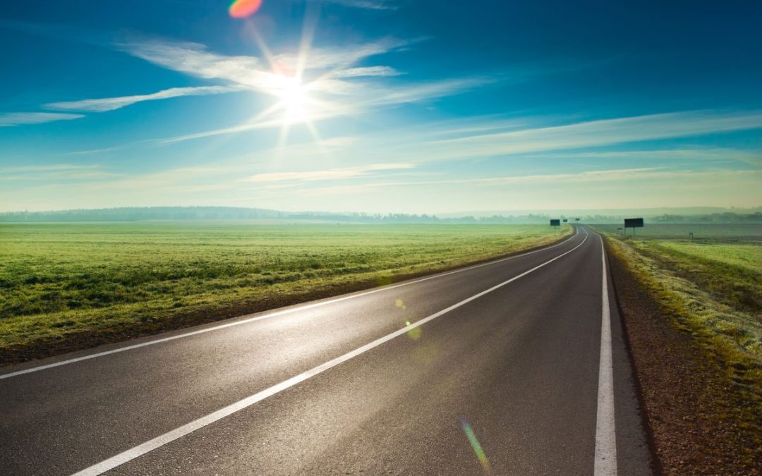 A view across flat open fields and an empty road with distant early morning mist and a bright shining sun