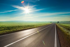 A view across flat open fields and an empty road with distant early morning mist and a bright shining sun