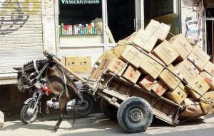 An overloaded trailer of goods packed into cardboard boxes tipped up leaving a donkey hanging in the air